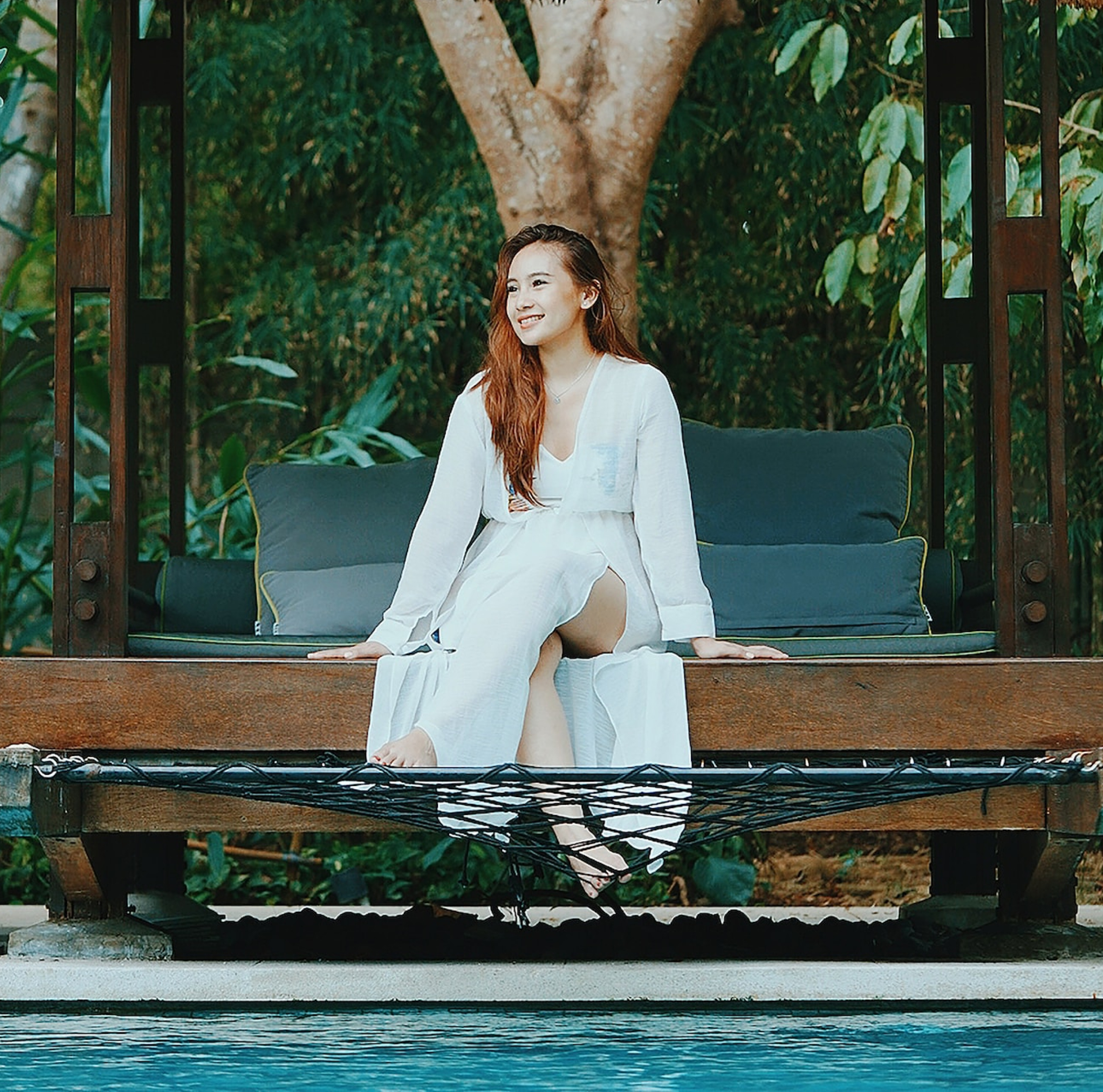 a member of a hospitality digital marketing agency with long brown and curly hair is sitting by the pool of a resort or hotel in bali wearing white pants, shirt, and cardigan