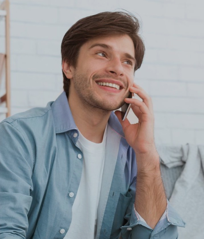 A brown-haired man wearing a simple white men's t-shirt and a blue denim shirt and long black denim pants is calling a friend while holding a debit or credit card about how to convert a customer into a conversion.